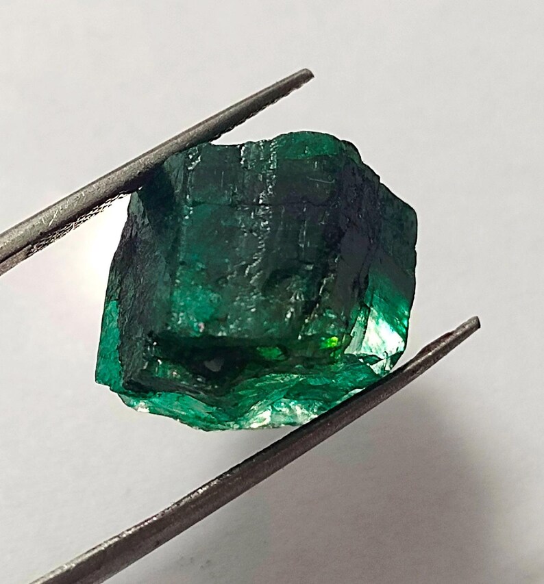 May include: A large, dark green emerald gemstone with a rough, faceted surface. The emerald is held in tweezers against a white background.