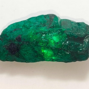 Può includere: Uno smeraldo grezzo non tagliato. La gemma è di un verde intenso e vibrante, con aree più scure e nere. La superficie ha una texture naturale e ruvida, con riflessi di luce.
