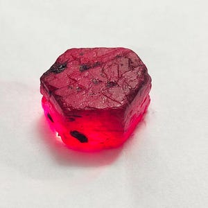 Puede incluir: Una gema de rubí en bruto, facetada, de color rojo intenso con inclusiones oscuras. La gema está iluminada desde abajo, creando un efecto brillante. La textura rugosa y la forma natural del rubí son visibles.