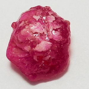 Puede incluir: Una piedra preciosa de rubí rosa en bruto con un aspecto translúcido y vítreo. El rubí tiene una forma natural irregular y mide aproximadamente 2,5 cm de diámetro.