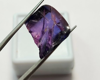 May include: A purple and blue gemstone with a faceted cut. The gemstone is held by tweezers and is in a white box.