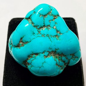 May include: A polished turquoise gemstone in a vibrant blue-green shade. The stone has a network of dark brown matrix veins across its surface. The gemstone is displayed on a black velvet surface.