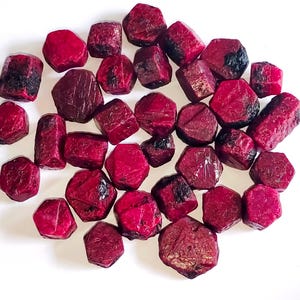 May include: A selection of raw ruby gemstones, varying in size and shape. The stones exhibit a deep red colour, with some areas of black. The gemstones are scattered on a white surface, highlighting their natural, rough texture.