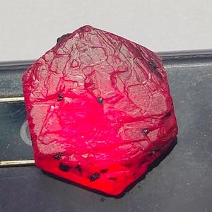Puede incluir: Una piedra preciosa de rubí rojo intenso con una superficie rugosa y facetada. La piedra preciosa tiene una forma hexagonal y mide aproximadamente 2,5 cm de diámetro.