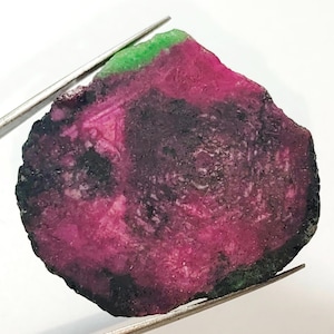May include: A rough, uncut gemstone with a green, pink, and black colour pattern. The gemstone is irregularly shaped and has a smooth, polished surface.