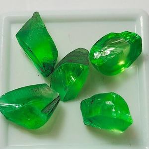 May include: Five green gemstones, cut and polished, with a translucent appearance. The gemstones are displayed on a white surface.