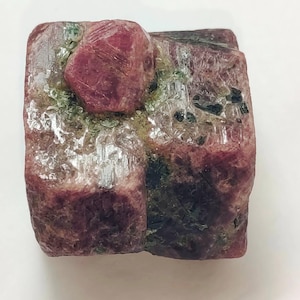 May include: A rough gemstone in shades of pink and red, with a prominent crystal formation. The stone has a textured surface with green and black inclusions. The overall shape is rectangular, with a natural, unpolished appearance. The gemstone is likely a mineral specimen.