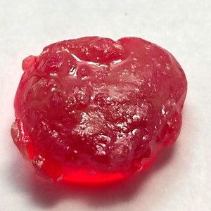Peut inclure: Une pierre précieuse rouge translucide de forme irrégulière. La surface présente un aspect texturé avec une finition légèrement brillante. La gemme est d'une couleur rouge profond et vibrante, avec des zones plus claires et plus foncées.