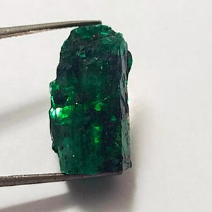 May include: A large, rough, green emerald gemstone. The emerald is a deep green color and has a slightly cloudy appearance. The emerald is being held by a pair of tweezers.