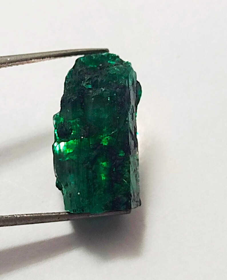 May include: A large, rough, green emerald gemstone. The emerald is a deep green color and has a slightly cloudy appearance. The emerald is being held by a pair of tweezers.