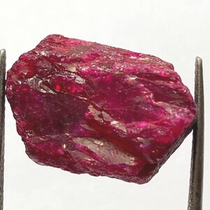 Könnte beinhalten: Ein rauer, ungeschliffener Rubin-Edelstein, gehalten von Metallpinzetten. Der Rubin hat eine tiefe, gesättigte rote Farbe mit einer leicht unebenen Oberfläche. Der Edelstein hat ein natürliches, rohes Aussehen.