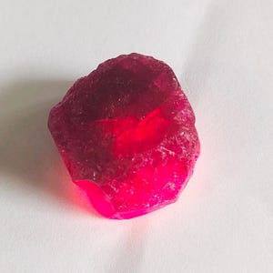 以下が含まれることがあります： 原石の天然ルビーの宝石。ルビーは深みのある鮮やかな赤色で、わずかに不規則な形をしています。表面は質感があり、ファセットと欠陥が見えます。