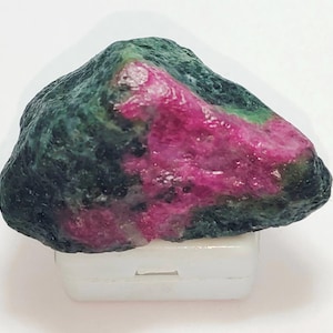 Peut inclure: Une pierre précieuse de rubis brute, non taillée, avec un extérieur vert foncé et un intérieur rose et rouge vif. La pierre, de forme irrégulière, repose sur une base blanche. Un ajout unique pour les collectionneurs de pierres précieuses.