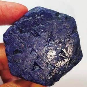 Puede incluir: Una piedra preciosa de zafiro azul oscuro en bruto con un patrón geométrico de líneas y triángulos. La piedra tiene un ligero brillo iridiscente.
