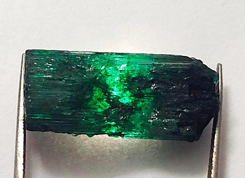 May include: A dark green emerald gemstone with a rough, striated surface. The emerald is translucent and has a slight black hue.
