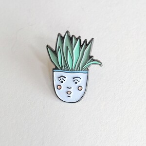 Pode incluir: Um broche de esmalte branco de uma planta em vaso com um rosto. A planta tem folhas verdes e o vaso tem uma borda azul clara. O rosto tem olhos pretos, um nariz rosa e uma boca rosa.