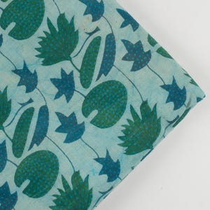 Puede incluir: Un trozo de tela doblado con un patrón repetitivo de nenúfares, hojas y tallos verde oscuro y turquesa sobre un fondo azul claro. La tela tiene un aspecto suave y ligeramente texturizado.