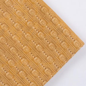 May include: A folded piece of fabric in a mustard yellow hue, featuring a repeating pattern of small, white floral and wavy designs. These elements are arranged in vertical rows. The fabric is angled against a white backdrop, showcasing its detailed texture and design.