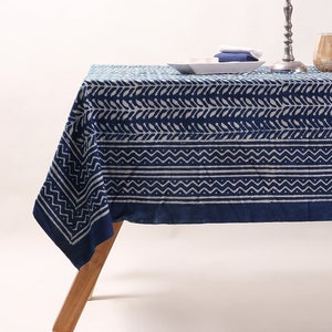 May include: A blue and white patterned tablecloth with a geometric design. The tablecloth is laid out on a wooden table with a silver candlestick and two glass vases with plants.