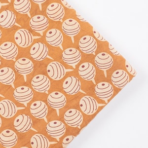 May include: Close-up of a fabric with a repeating pattern of cream-coloured, striped ball shapes on a tan background. The balls have a small stem and are evenly spaced across the fabric. The fabric is folded over on a white surface.