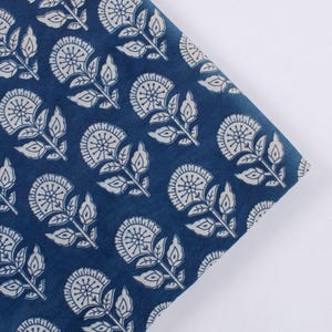 May include: Close-up of folded fabric with a repeating floral pattern. The fabric is a deep indigo blue, and the floral design is white. The flowers have a circular shape with detailed petals and are connected by stems and leaves.