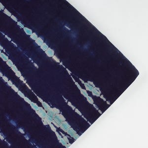 Könnte beinhalten: Dunkelblauer Stoff mit einem Batikmuster mit hellblauen und weißen diagonalen Streifen. Der Stoff ist gefaltet und zeigt die Textur und das Design des Materials. Der Hintergrund ist schlicht weiß.