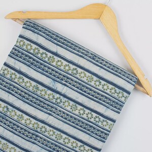 May include: A folded fabric with a blue and white striped pattern, accented with rows of small green floral designs. The fabric is hanging on a wooden hanger, showcasing its texture and design. The fabric appears to be cotton or a similar material.