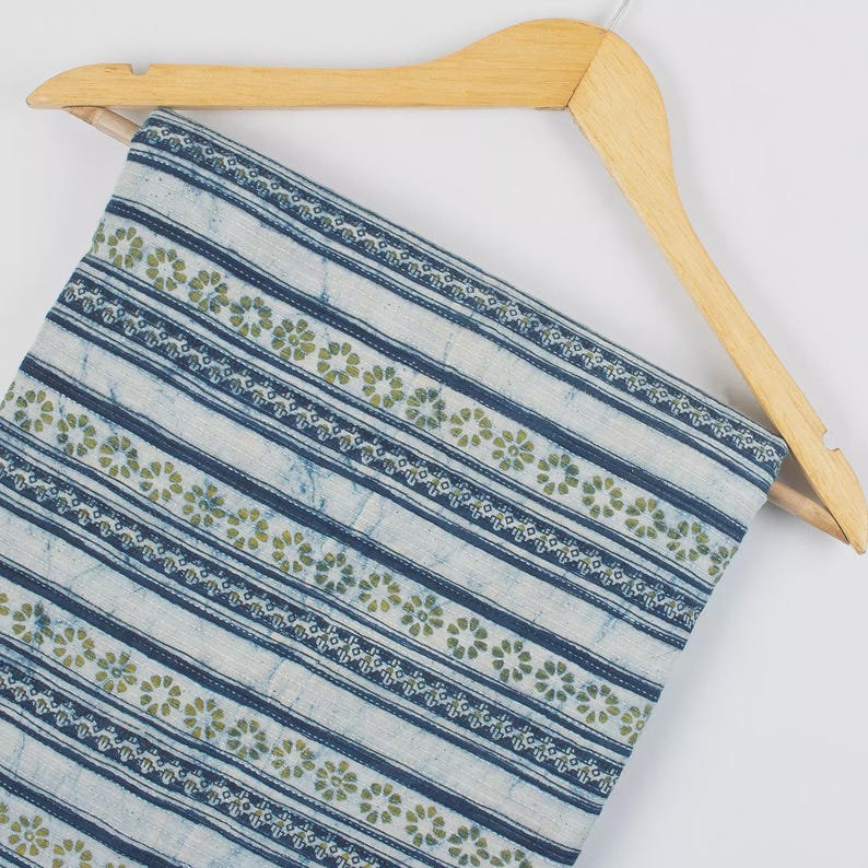 May include: A folded fabric with a blue and white striped pattern, accented with rows of small green floral designs. The fabric is hanging on a wooden hanger, showcasing its texture and design. The fabric appears to be cotton or a similar material.