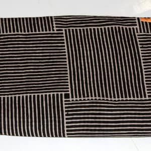 May include: Folded fabric with a geometric pattern of black vertical and horizontal stripes on a tan background. The fabric is rectangular and appears to be cotton or a similar material. A small orange tag is visible in the upper right corner.