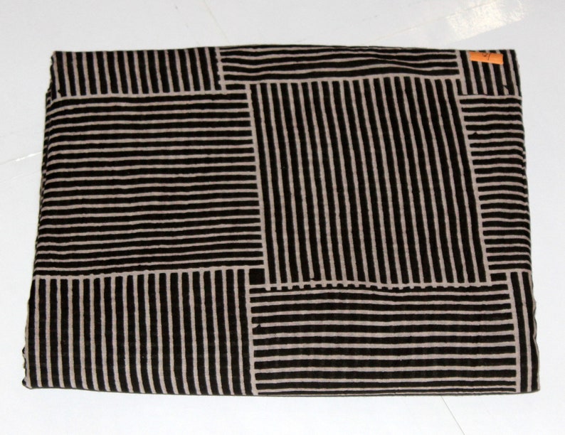 May include: Folded fabric with a geometric pattern of black vertical and horizontal stripes on a tan background. The fabric is rectangular and appears to be cotton or a similar material. A small orange tag is visible in the upper right corner.