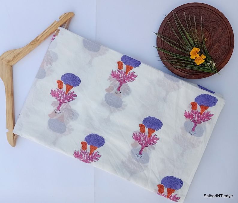 Puede incluir: Tela blanca con un estampado floral repetido en morado, naranja y rosa. La tela est&aacute; doblada y colocada sobre una percha de madera. Un plato de madera con flores y vegetaci&oacute;n est&aacute; en el fondo.