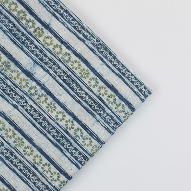 May include: A folded piece of fabric with a striped pattern. Alternating stripes of white and navy blue, with a floral design in green. The fabric has a soft, textured appearance, suitable for various textile projects.