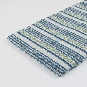 May include: Folded fabric with a repeating pattern of blue stripes and floral motifs in green and white. The fabric appears to be cotton or a similar material, with a slightly textured surface. The design is printed on a white background.