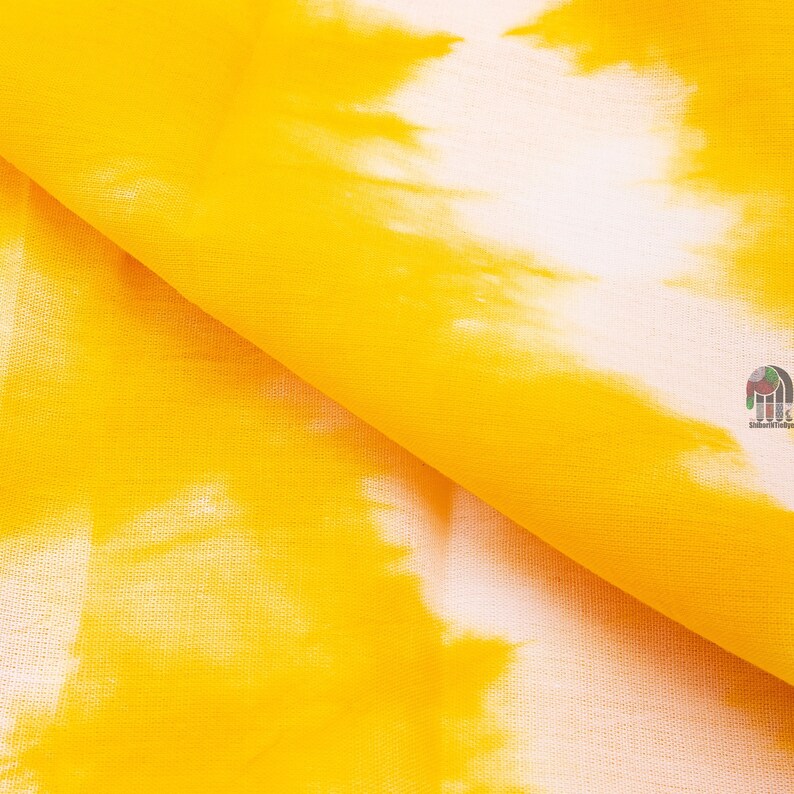May include: Yellow fabric with a white tie-dye pattern. The fabric is folded in half, showing the texture of the material.