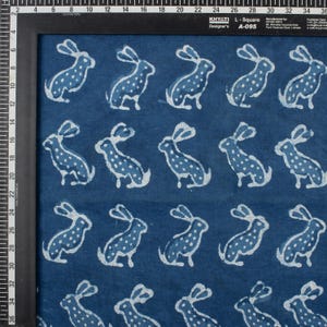 May include: Indigo blue fabric with a repeating pattern of white rabbits. Each rabbit is depicted in a seated position with long ears and dotted details. The fabric is framed by a black border.