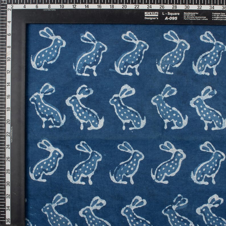 May include: Indigo blue fabric with a repeating pattern of white rabbits. Each rabbit is depicted in a seated position with long ears and dotted details. The fabric is framed by a black border.