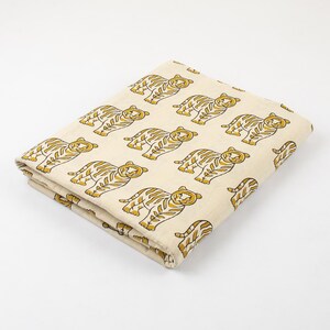 May include: Folded fabric with a repeating pattern of yellow tigers on a cream background. The tigers have detailed stripes and facial features. The fabric is neatly folded, showcasing the design's repeat.