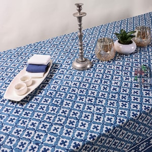 Puede incluir: Un mantel de mesa con estampado azul y blanco con un diseño geométrico. El mantel está puesto con un candelero de plata, dos cuencos pequeños blancos y una bandeja rectangular blanca para servir con dos toallas azules y blancas. También hay una pequeña planta en maceta y dos portavelas de vidrio transparente.