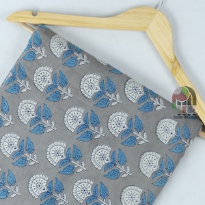 Pu&ograve; includere: Tessuto di cotone grigio con stampa floreale bianca e blu. Il tessuto &egrave; piegato e drappeggiato su un appendiabiti in legno.