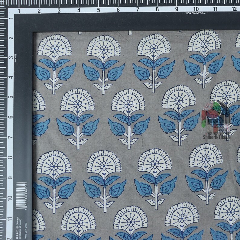 Pu&ograve; includere: Primo piano di un tessuto di cotone grigio con un motivo floreale bianco e blu. Il tessuto &egrave; stampato con un motivo ripetitivo di fiori stilizzati con foglie blu e centri bianchi.