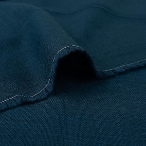 Puede incluir: Primer plano de un trozo de tela verde azulado doblada. La tela tiene un tejido texturizado y un borde sin rematar con costuras blancas. El color es un verde azulado profundo y saturado, con textura y pliegues visibles.