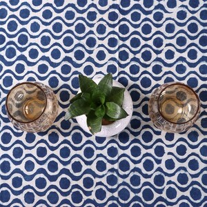 May include: Two brown glass candle holders and a potted green plant with white ceramic pot on a white and blue patterned tablecloth.