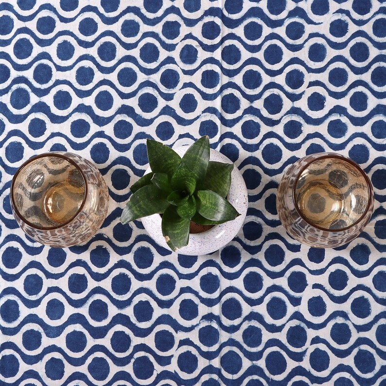 May include: Two brown glass candle holders and a potted green plant with white ceramic pot on a white and blue patterned tablecloth.