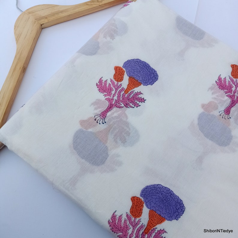 Puede incluir: Tela blanca con un estampado floral repetido. Las flores son moradas, naranjas y rosas, con hojas verdes. La tela est&aacute; doblada y colgada de una percha de madera.