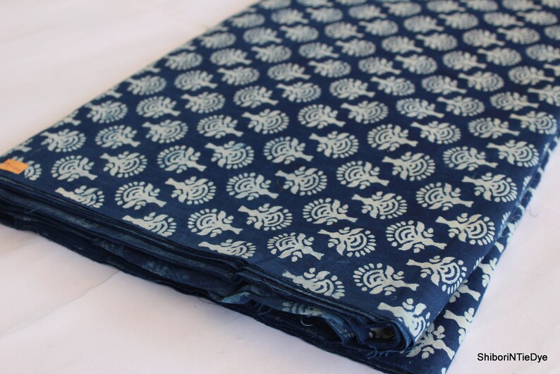 May include: A folded stack of indigo blue cotton fabric with a white floral pattern. The fabric is folded in thirds and has a white tag with the text "ShiboriNTieDye" on the bottom right corner.