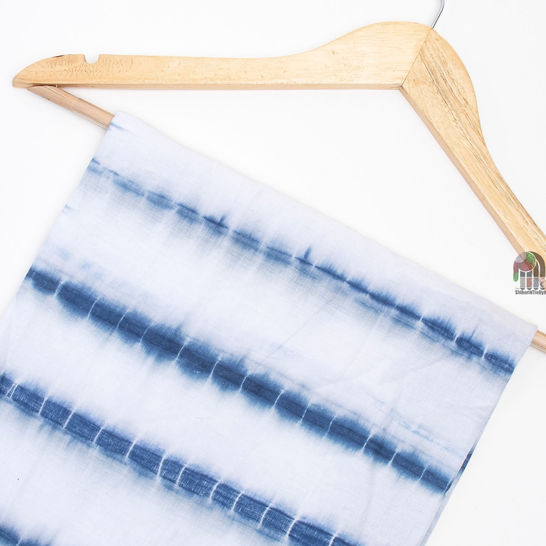 Op de afbeelding: Een witte stof met blauwe tie-dye strepen hangend aan een houten hanger. De stof heeft een zachte, vloeiende textuur.