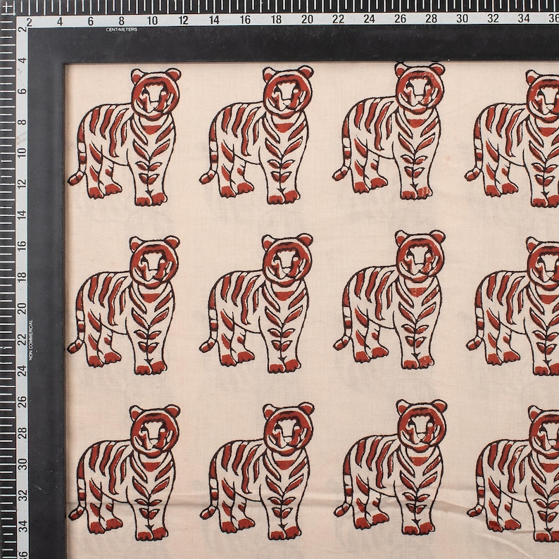 May include: Cream-colored fabric featuring a repeating pattern of stylized tigers. Each tiger is depicted in a standing pose with red-brown stripes and details. The fabric is framed with a black border and includes a centimeter ruler.