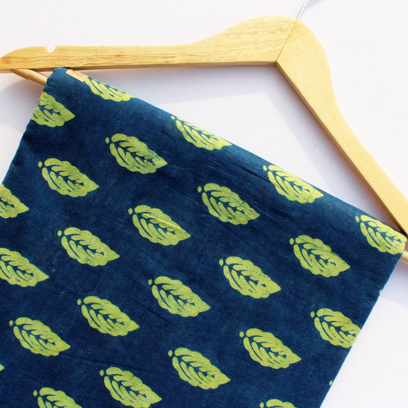 Op de afbeelding: Een blauwe stof met een herhalend patroon van groene bladeren. De stof hangt aan een houten hanger.