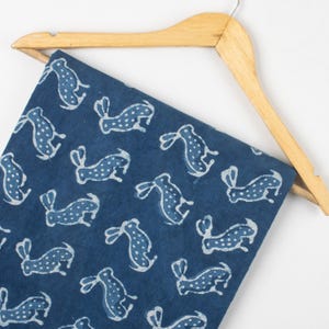 May include: A navy blue fabric with a repeating pattern of white rabbit silhouettes. The fabric is draped over a wooden hanger, showcasing the design and texture. The rabbits have dotted bodies and long ears.