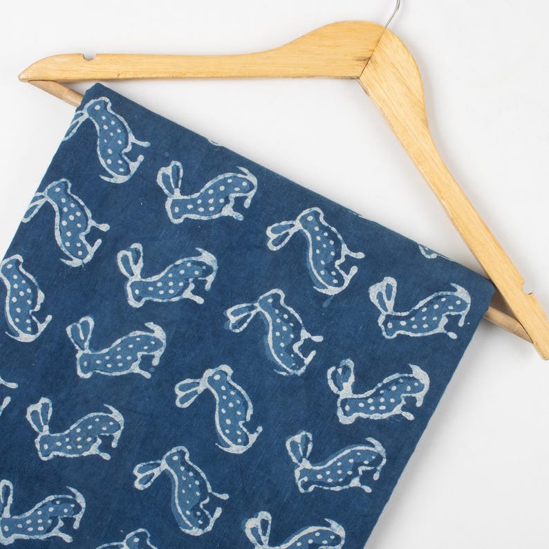 May include: A navy blue fabric with a repeating pattern of white rabbit silhouettes. The fabric is draped over a wooden hanger, showcasing the design and texture. The rabbits have dotted bodies and long ears.